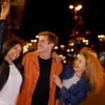Three young people wave at the camera while standing together on a lively street